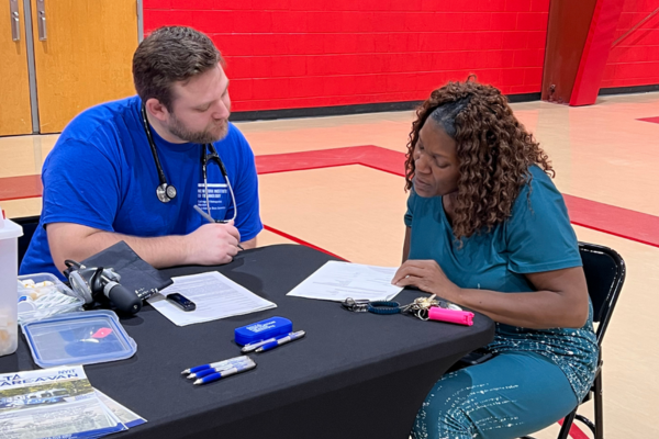 A healthcare worker in a blue shirt talks to a woman in green. They sit at a table with papers in a gym and are focused.
