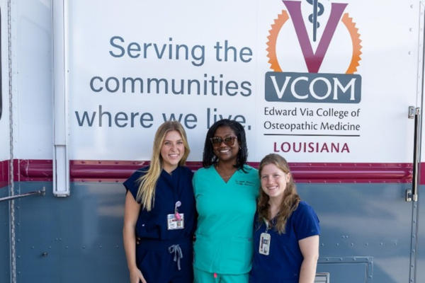 Three people in medical scrubs smiling in front of a VCOM mobile unit. The sign reads