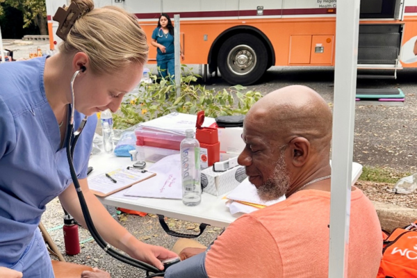 A clinician checks a man's blood pressure at an outdoor community event. Behind them is a mobile clinic van labeled