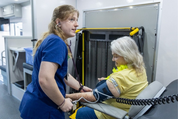 A healthcare worker in blue scrubs checks the blood pressure of an older woman in a yellow shirt. They are inside a medical facility. The mood is professional and caring.