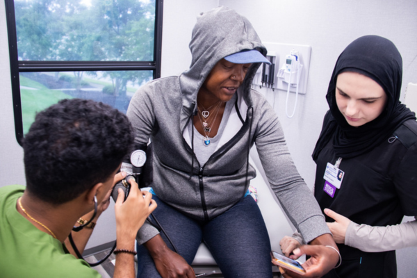 A patient in a gray hoodie gets their blood pressure checked by a healthcare worker in a medical clinic. Another clinician stands nearby, assisting. Caption: VCOM-Carolinas students deliver care to patients at an MMU event.