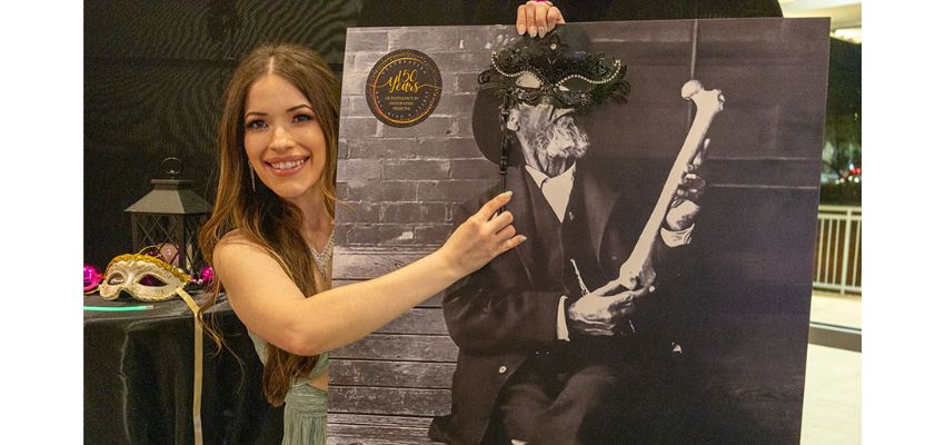 A young woman poses next to a large historical-themed poster, holding up a decorative mask.