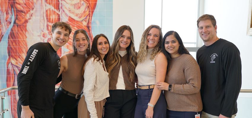 A group of medical students from the Idaho College of Osteopathic Medicine (ICOM) pose together, smiling in front of an anatomical mural.