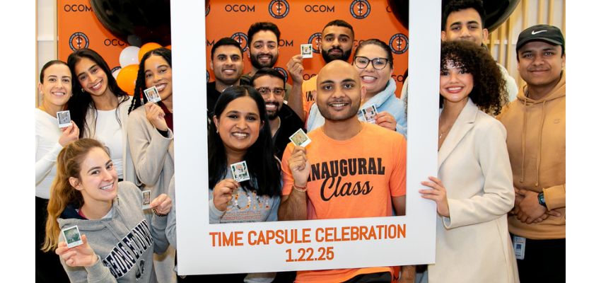 A group of students from the Oklahoma College of Osteopathic Medicine (OCOM) celebrate their time capsule event, holding up small mementos with a branded backdrop.