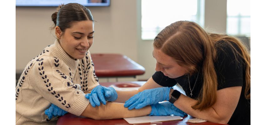 A medical student at VCOM Louisiana practices a clinical skill on a fellow student, who is wearing gloves and using a needle or similar instrument.