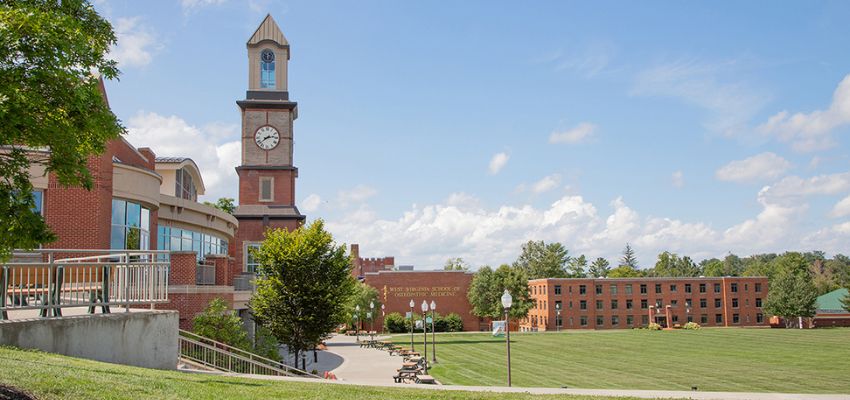 A scenic view of the West Virginia School of Osteopathic Medicine (WVSOM) campus, featuring a clock tower, academic buildings, and a large green lawn under a blue sky.