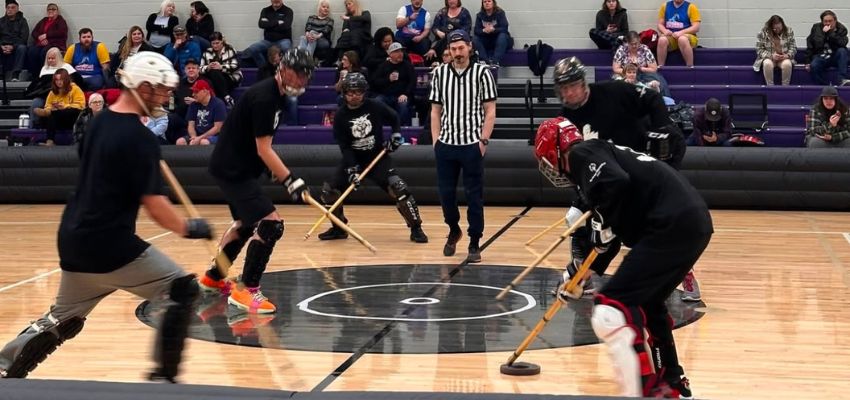 A group of players in protective gear compete in a broomball game on an indoor court while a referee watches.