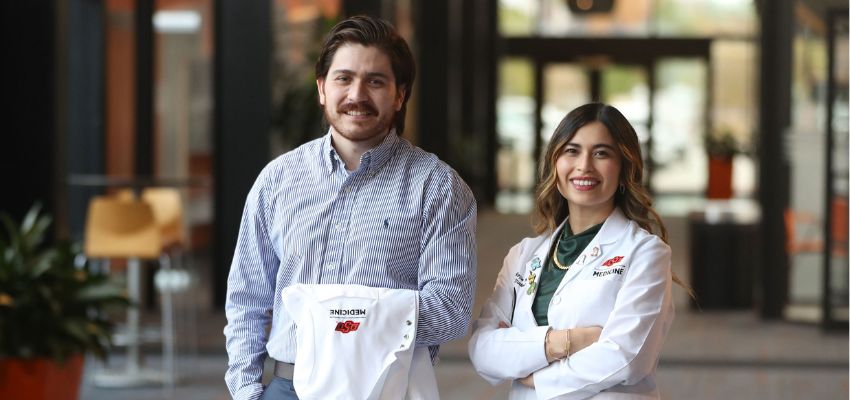 Two medical students stand together, one holding a white coat, in a modern indoor setting.