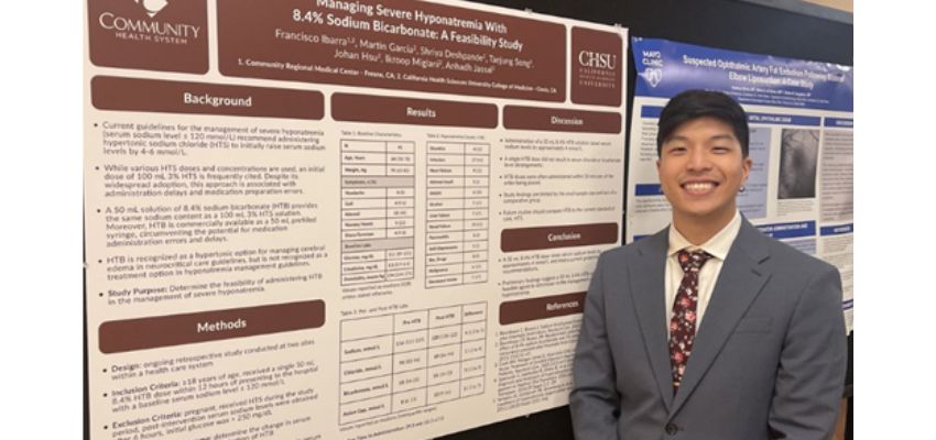 A young man in a suit stands smiling in front of a research poster presentation, representing CHSU-COM.