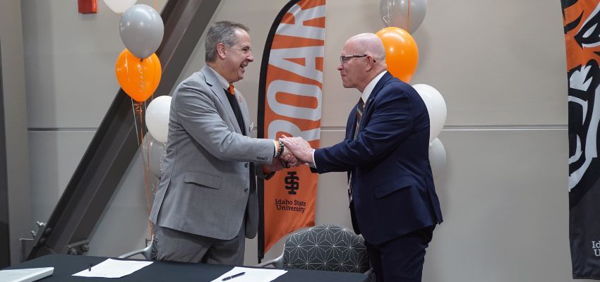 Two men in suits shake hands at a signing event with Idaho State University banners and balloons in the background.