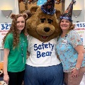 TwowomenposewithapersoninabearmascotcostumewearingaSafetyBearshirtinfrontofawallfilledwithteddybears