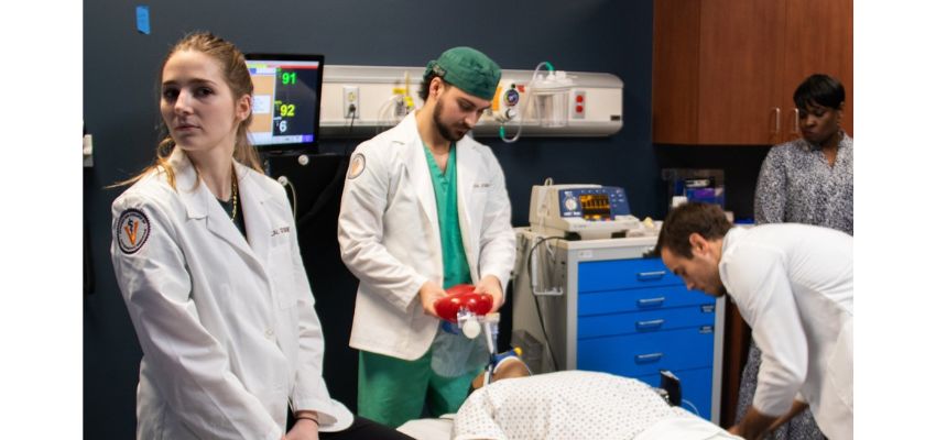 Medical students in white coats participate in a hands-on clinical training exercise in a simulated hospital setting.