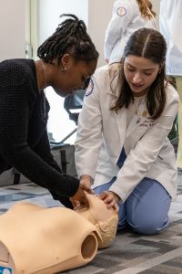 A medical student in a white coat guides another person through a CPR demonstration on a mannequin.