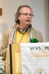 A speaker in a yellow sweater and striped scarf stands at a podium with a 