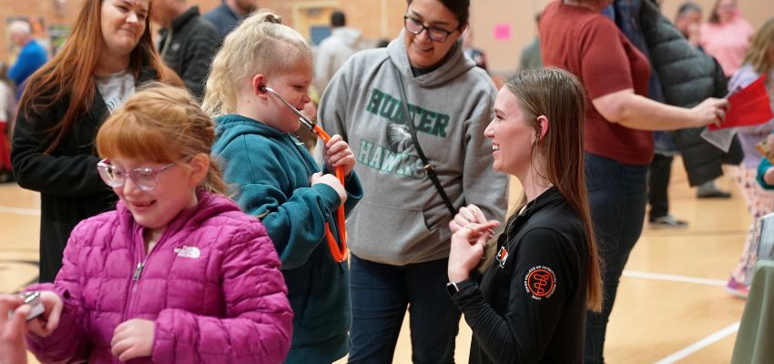 A young girl uses a stethoscope while interacting with a smiling medical student in a black jacket at a community event.