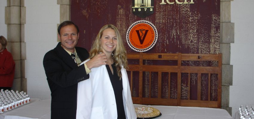 A father in a suit places a white coat on his smiling daughter during a ceremony, with a VCOM banner in the background.