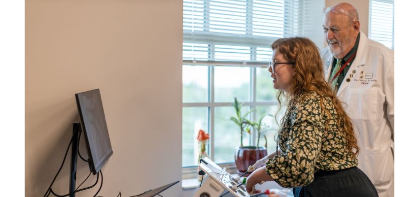 A student uses medical equipment while an older faculty member in a white coat observes in a classroom setting.