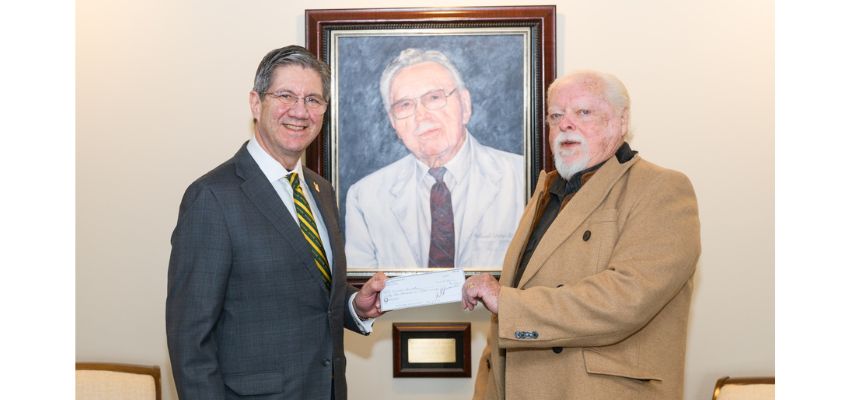 Two men in suits shake hands while exchanging a check, standing in front of a portrait of a distinguished doctor.