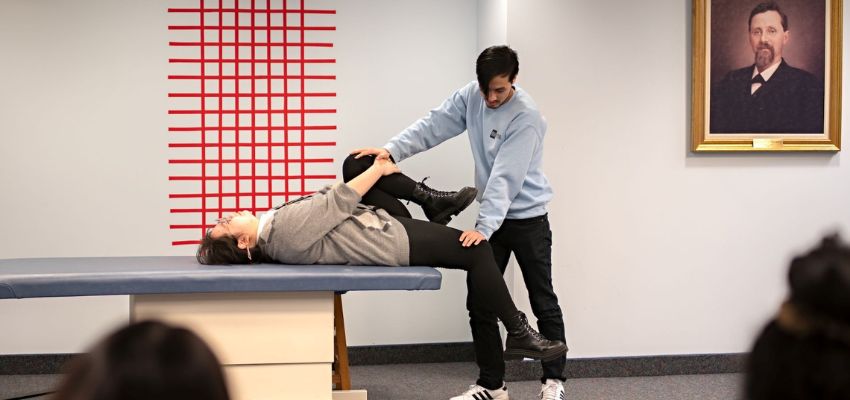 A student in a light blue sweatshirt performs a hands-on osteopathic technique on a peer lying on a treatment table, with a historical portrait hanging on the wall.