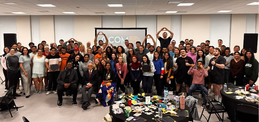 A large group of students gathers for a group photo at a conference or workshop, with some forming hand gestures above their heads.