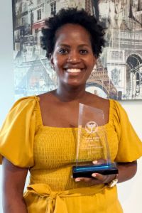 A woman in a yellow dress holds a glass award and smiles proudly in front of a cityscape-themed wall.