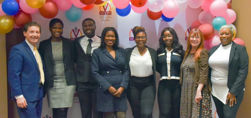 A diverse group of eight individuals stands together under colorful balloons in front of a VCOM Virginia backdrop, smiling for a group photo.