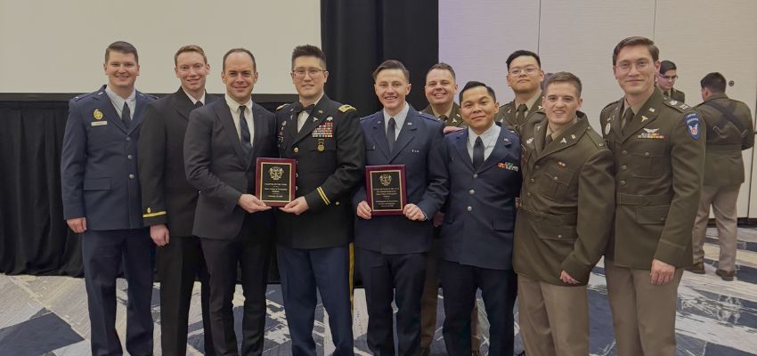 A group of military service members and students in dress uniforms pose together at an awards ceremony, with two individuals holding plaques.