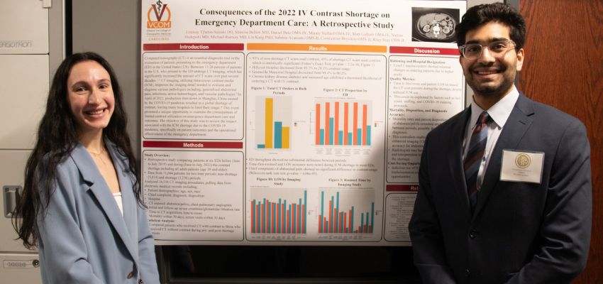 Two students stand beside a research poster presentation on the impact of the 2022 IV contrast shortage in emergency departments, smiling at the camera.