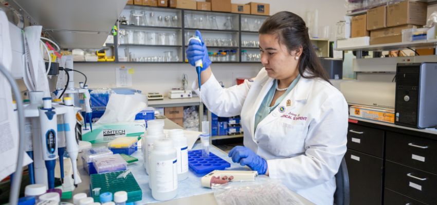 A medical student wearing a lab coat and gloves carefully uses a pipette while conducting research in a laboratory setting.