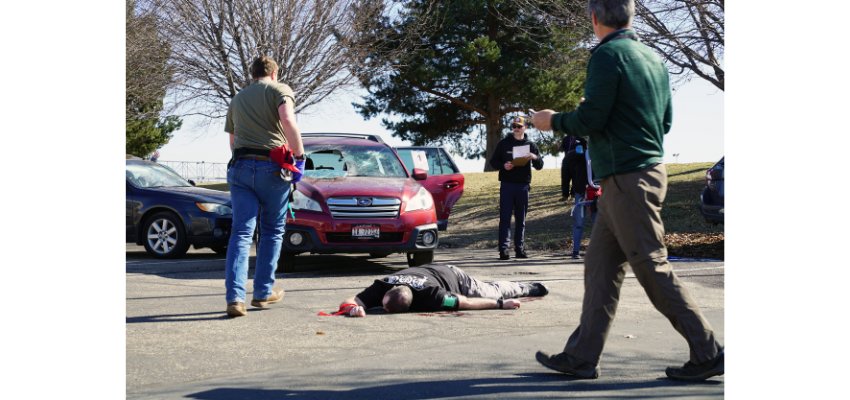 A simulated emergency training scenario takes place in a parking lot, with a person lying on the ground while others respond.