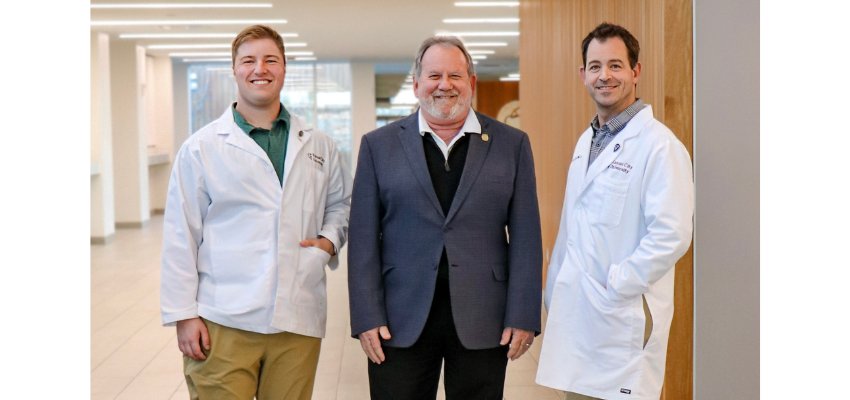 A medical student in white coats stands beside two faculty members, smiling inside a modern academic building.