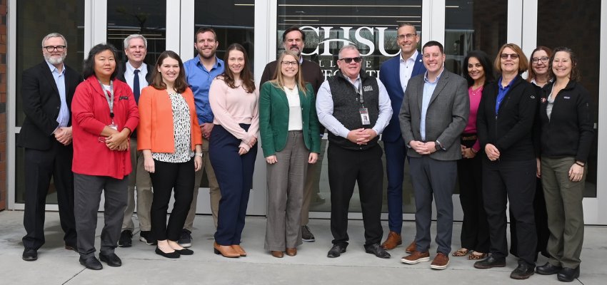 A group of faculty and staff pose in front of a CHSU College of Osteopathic Medicine sign, dressed in business and casual attire.