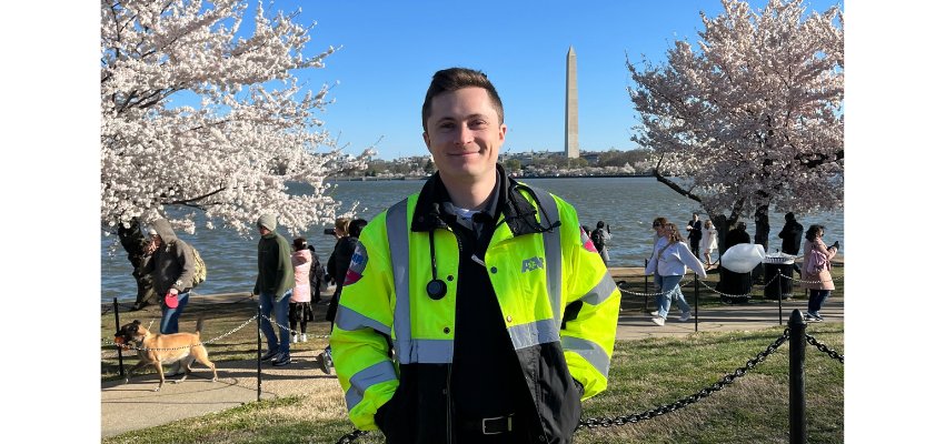 A young medical professional wearing a neon yellow EMS jacket stands in front of the Washington Monument and cherry blossoms, smiling at the camera.