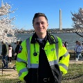 AyoungmedicalprofessionalwearinganeonyellowEMSjacketstandsinfrontoftheWashingtonMonumentandcherryblossomssmilingatthecamera