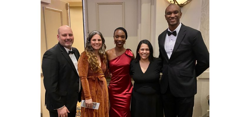 A group of five individuals dressed in formal attire poses at a gala event, smiling for the camera.