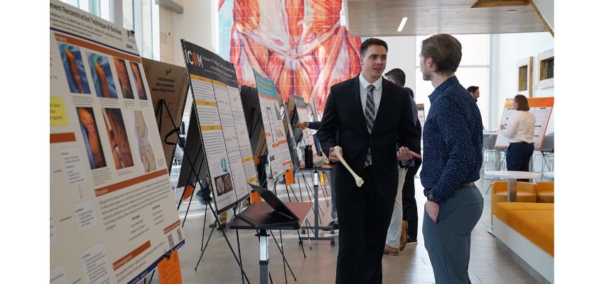 A student discusses a research poster with a peer during a poster session in a bright, modern academic building.