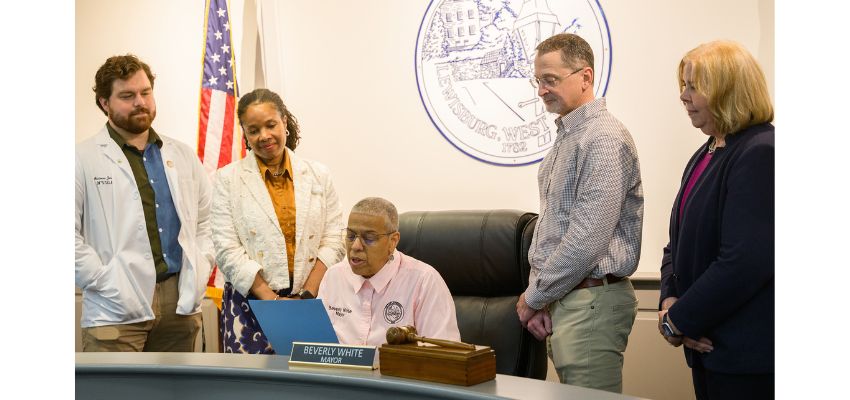 A group of individuals, including medical professionals, gathers around the mayor of Lewisburg, West Virginia, during a ceremonial event at City Hall.