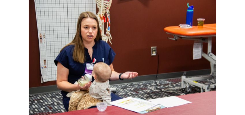 A woman in scrubs sits holding an infant and a bottle, demonstrating or discussing infant care in a clinical or educational setting.