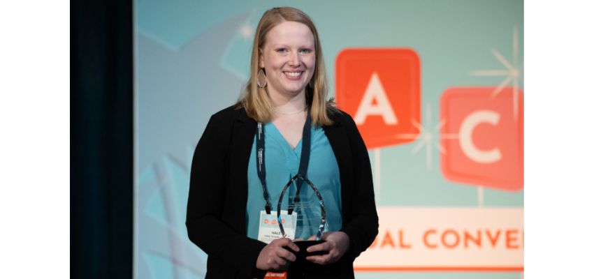 A woman smiles while holding an award on stage at a professional conference with a backdrop.