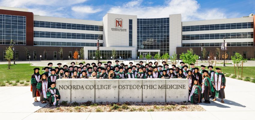 A large group of graduates pose in regalia in front of the Noorda College of Osteopathic Medicine building.