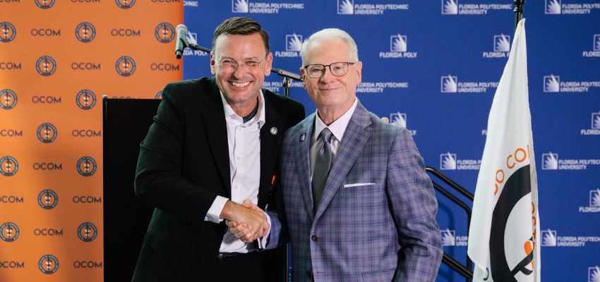 Two men in suits shake hands in front of a backdrop displaying OCOM and Florida Polytechnic University logos.