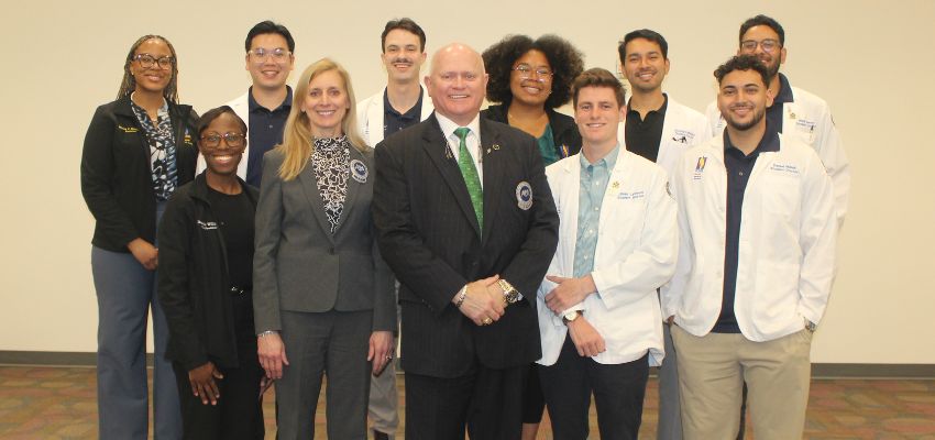 A group of medical students and faculty, some in white coats, pose for a photo together in a classroom setting.