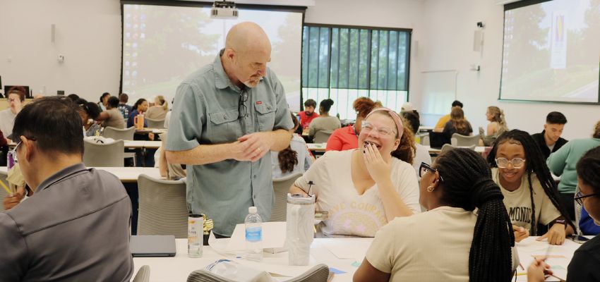 A classroom of students work in groups while a faculty member engages with one table during an interactive session.