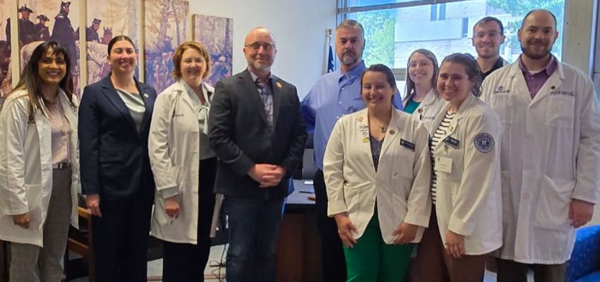 A group of medical students and professionals pose for a photo inside an office, some wearing white coats.