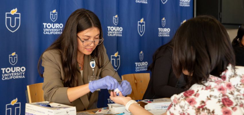 A student wearing gloves performs a finger-prick test on someone’s hand at a Touro University California event table.