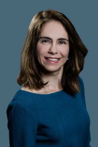 A woman in a blue dress smiles in a professional headshot against a solid background.