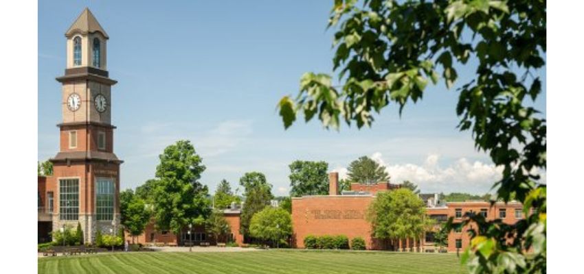 A scenic view of a campus shows a tall clock tower, brick buildings, and a large green lawn framed by tree branches.