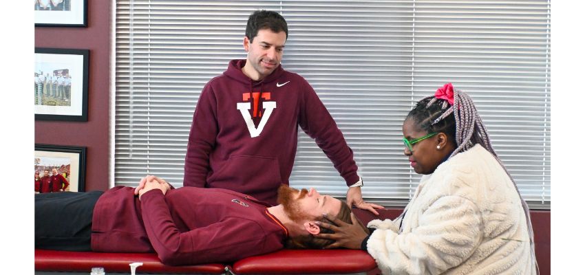 Students practice osteopathic manipulative treatment as one student lies on a table while two others observe and assist in a classroom setting.