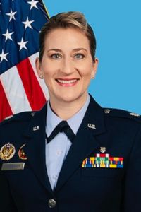 A woman in a military uniform smiles in a formal portrait with an American flag in the background.