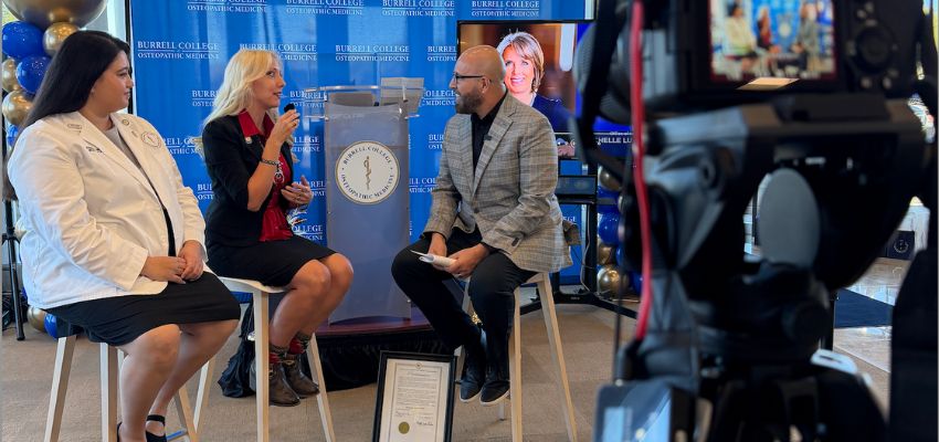 Three people sit on stools participating in a filmed discussion at a Burrell College of Osteopathic Medicine event, with a camera visible in the foreground.